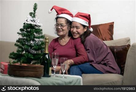 Two 60s old mother and daughter wearing santa hat, hug and sitting in living room at home. Christmas or New Year Celebration Concept.