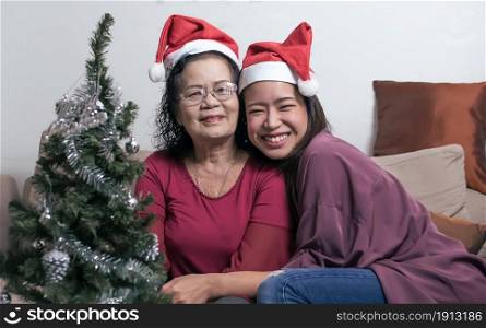 Two 60s old mother and daughter wearing santa hat, hug and sitting in living room at home with blur foreground of tree and bottle. Christmas or New Year Celebration Concept.
