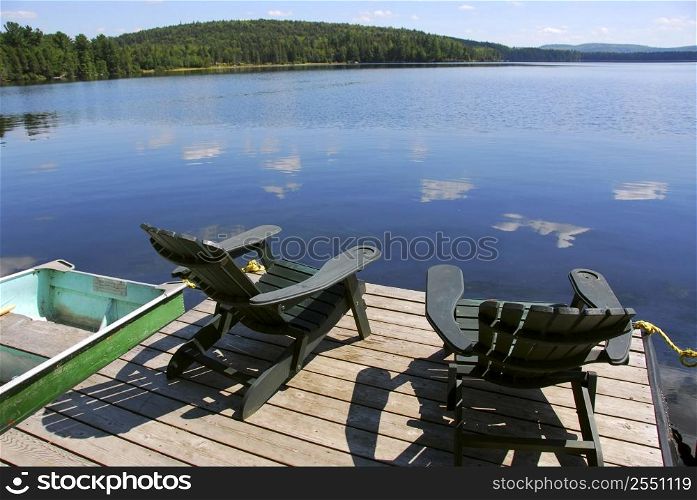Two adirondack wooden chairs on dock facing a blue lake with clouds reflections