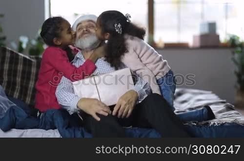 Two adorable affectionate mixed race granddaughters kissing beloved grandfather in santa hat with christmas gift box on lap on the cheeks at christmas. Cute girls giving a kiss to joyful grandpa and falling on bed in love embrace on winter holidays.