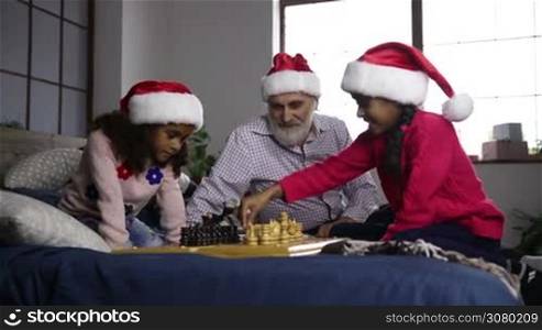 Two adorable african american sisters in santa hats playing chess on bed during family spending christmas time at home. Excited little mixed race girl giving high five to her caucasian grandfather while playing chess with younger sister on Xmas eve.
