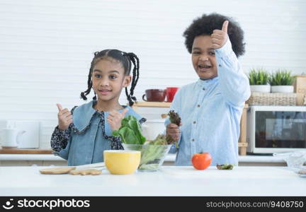 Two African cute sister and brother smiling, thumbs up cheering for eat fresh vegetables at home kitchen. Boy holding green salad vegetable in hand. Healthy food, good for kids