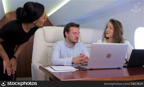 Two analysts working on laptop on board of private jet. Biracial flight attendant offer her service to caucasian businessman and businesswoman traveling inside of business airplane cabin.. Two businessmen work on private plane
