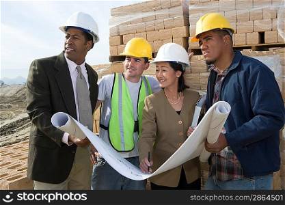 Two architects and two construction workers standing on construction site holding blueprints