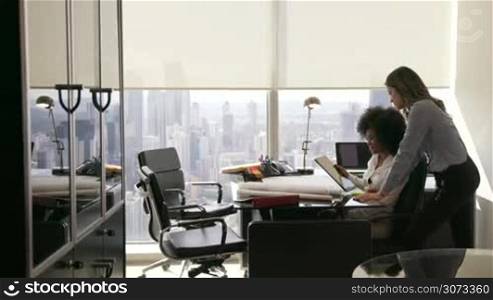 Two architects in modern office building, sitting on desk with blueprints and housing projects. The women hold a tablet and surf the web. They smile and talk each other. Wide shot