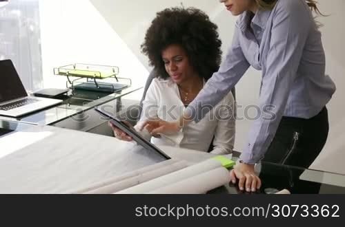 Two architects in modern office building, sitting on desk with blueprints and housing projects. The women hold a tablet and surf the web. They smile and talk each other. Medium shot