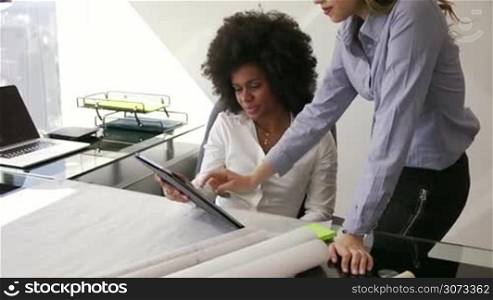 Two architects in modern office building, sitting on desk with blueprints and housing projects. The women hold a tablet and surf the web. They smile and talk each other. Medium shot