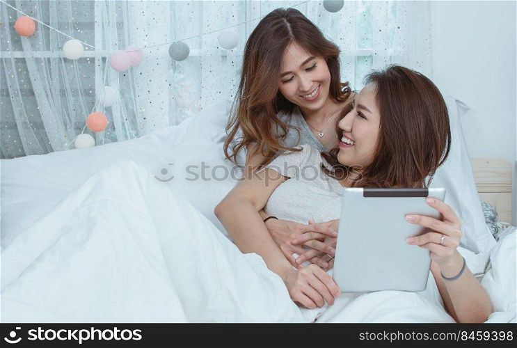Two asian beautiful women laying down on cozy bed, hug and using tablet together. Lifestyle and LGBT Concept.