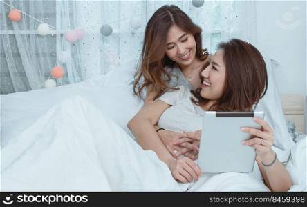 Two asian beautiful women laying down on cozy bed, hug and using tablet together. Lifestyle and LGBT Concept.
