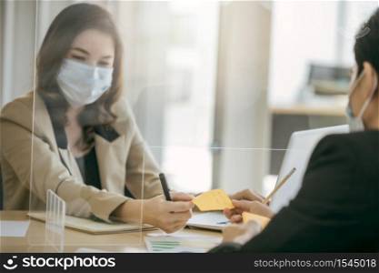 Two Asian business office wearing using partition to social distancing for protect coronavirus covid-19 pandemic. Social and business distancing new normal lifestyle.