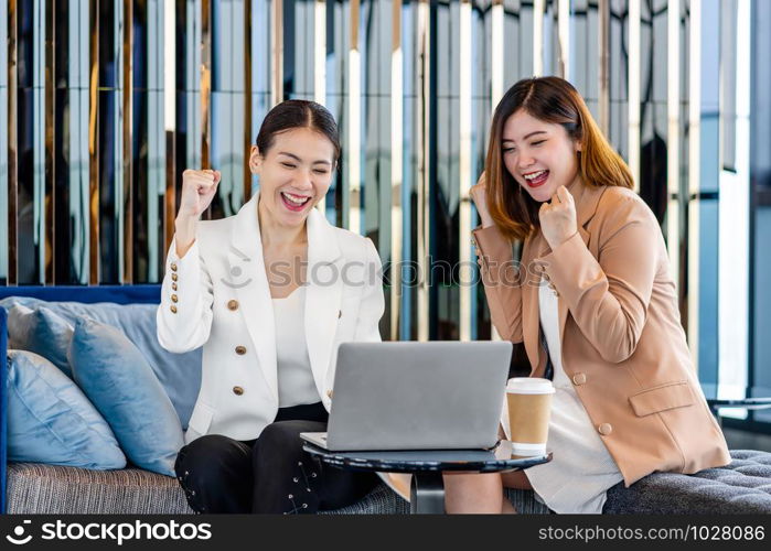 Two asian businesswomen hapiness celebrating together when checking success goal via technology laptop in modern office or coworking space, coffee break, relaxing and talking after working time