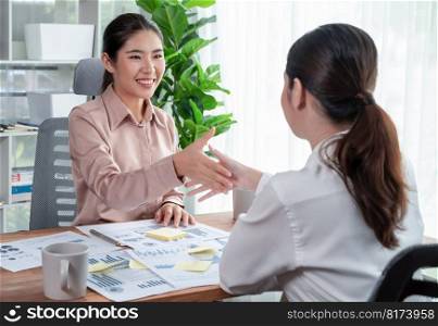 Two Asian businesswomen shaking hands in modern office after successfully analyzing piles of dashboard data papers, as importance of teamwork and integrity in the workplace concept. Enthusiastic. Two Asian businesswomen handshake in modern office with data paper. Enthusiastic