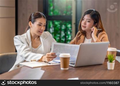 Two asian businesswomen working with the partner business with paper graph document and technology laptop in modern meeting room, office or working space, coffee break, partner and colleague concept