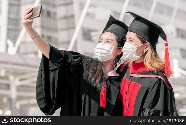 Two asian female students wearing face mask to protect virus and using mobile phone to take selfie. Graduation, Education, Technology and Health Concept.