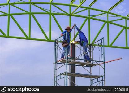 Two Asian welders on scaffolding are welding metal roof structure of industrial building against blue sky background