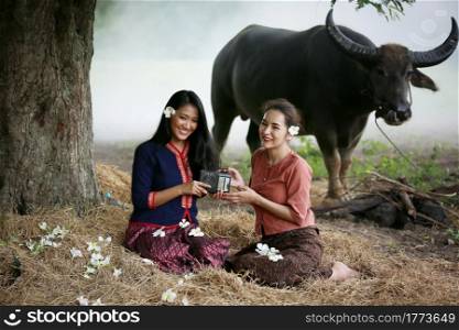 Two Asian woman wearing traditional thai Esan custom style culture sitting in field, while listening vintage style radio against buffalo and farm background.