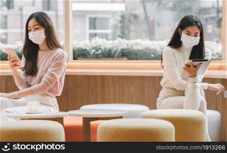 Two Asian women wearing mask to protect virus and keeping social distancing while using mobile phone and tablet in coffee shop . New Normal Concept.