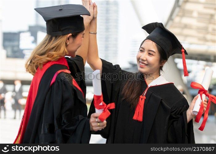 Two Asian young beautiful graduate fema≤students with master and bachelor°ree smiling and holding diploma in hand and Congratulations with hi five hand. Blur background of University building