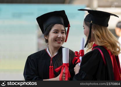 Two Asian young beautiful graduate female students with master and bachelor degree standing smiling and holding diploma in hand expressing proud face. Blur background of University building