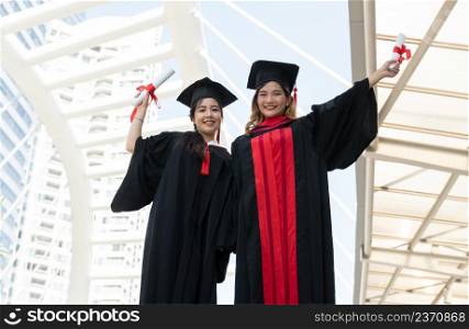 Two Asian young beautiful graduate female students with master and bachelor degree standing smiling and holding diploma in hand expressing proud face. Blur background of University building