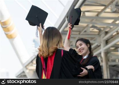Two Asian young beautiful graduate female students with master and bachelor degree holding black board cap with red tassels up to the sky is smiling. Blur background of University building
