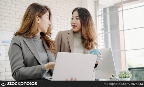 Two Asian young creative businesswomen holding a cup of coffee, working on laptop and disscuss about work while working in office. Casual business, freelance work at cafe, social meeting concept.