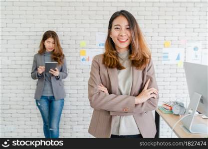 Two Asian young creative businesswomen holding a cup of coffee, working on laptop and disscuss about work while working in office. Casual business, freelance work at cafe, social meeting concept.