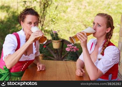 Two attractive Bavarian women wearing traditional dirndl dresses sitting at an outdoor table drinking beer from long glasses during the celebrations for the Oktoberfest