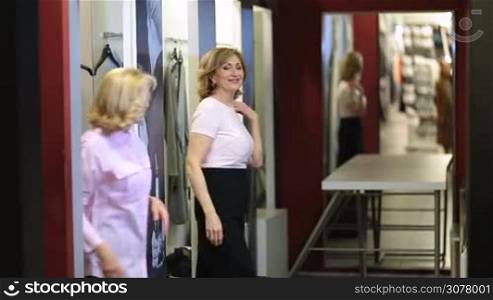 Two attractive female friends trying on clothes in fashion store fitting room. Beautiful senior women shopping together at garments shop, choosing new apparels, showing new clothing to each other and looking at themselves in the mirror.