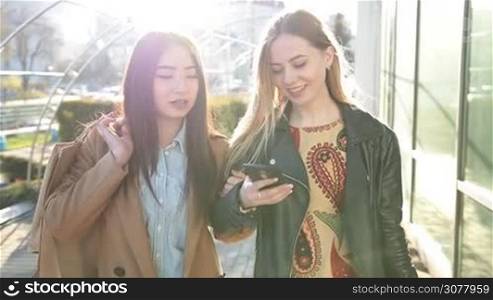 Two attractive multiethnic girl friends with shopping bags using smart phone while walking in city street after shopping in glow of amazing sunset. Slow motion. Females shopaholics making online purchase with mobile phone outdoors.