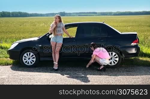 Two attractive women on high heels stopped on a roadside of a rural road during roadtrip because tyre is punched. Cute girl is lifting up car using screw-jack to change flat tire with spare one.