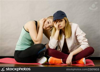 Two attractive women wearing sports clothes sitting on exercise mat being bored or tired after hard workout.. Two sporty women being bored