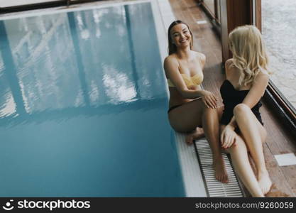Two attractive young women in bikini sits on the poolside of the indoor swimming pool