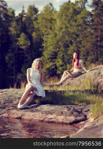 Two attractive young women sits on the rock, summer day on the lake