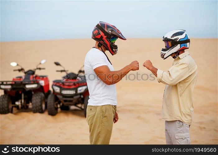 Two atv racers in helmets banging their fists before freedom riding in desert. Male persons on quad bikes, sandy race, dune safari in hot sunny day, 4x4 extreme adventure, quad-biking. Two atv racers in helmets banging their fists