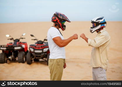 Two atv racers in helmets banging their fists before freedom riding in desert. Male persons on quad bikes, sandy race, dune safari in hot sunny day, 4x4 extreme adventure, quad-biking. Two atv racers in helmets banging their fists