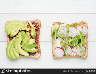 Two avocado toasts with mashed avocado and beetroot,sliced avocado ,radish, snow pea sprouts and goat cheese on white wooden background