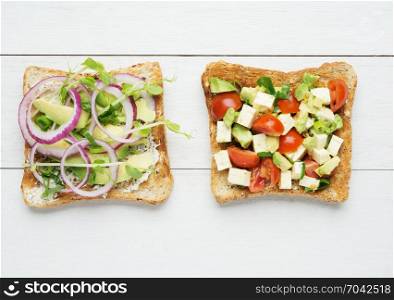 Two avocado toasts with mashed avocado and onion,sliced avocado ,tomato, snow pea sprouts and goat cheese on white wooden background
