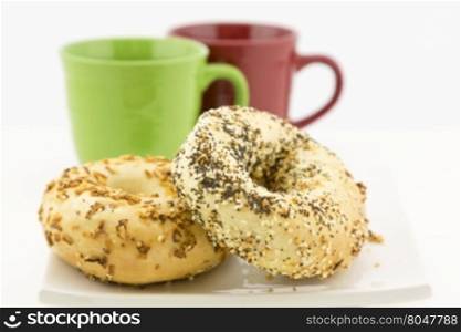 Two bagels on white plate with red and green mugs behind. Selective focus on breakfast bread.