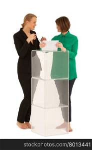Two barefoot women looking to each other while talking near stand made of glass cubes. One of them holding tablet. White background. Isolated. Two women looking to each other while talking