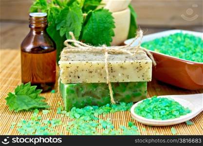 Two bars of homemade soap with twine, bath salt, oil bottle, nettle in a mortar on a wooden boards background