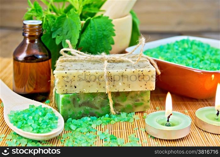 Two bars of homemade soap with twine, bath salt, oil bottle, nettle in a mortar, scented candle on a wooden boards background