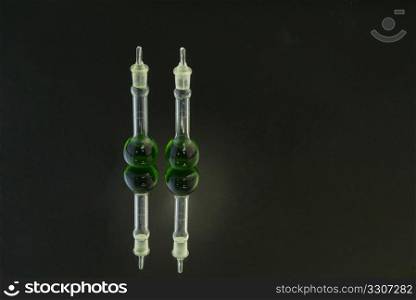 Two beakers with green fluid on mirror surface against black background with lighting highlight on beakers; copy space;