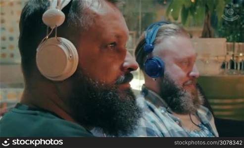 Two bearded men relaxing on the sofa in trade centre and listening to music in headphones