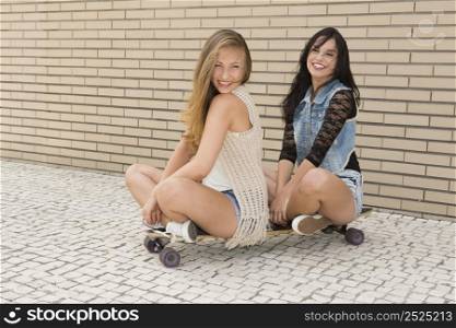 Two beautiful and young girlfriends having fun with a skateboard, in front of a brick wall