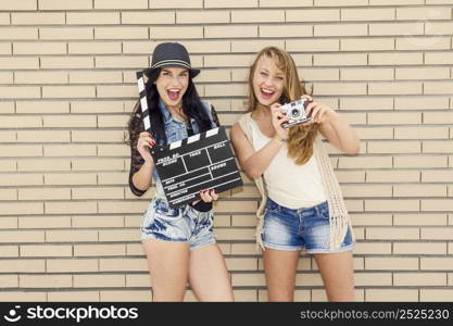 Two beautiful and young girlfriends holding a clapboard, in front of a brick wall