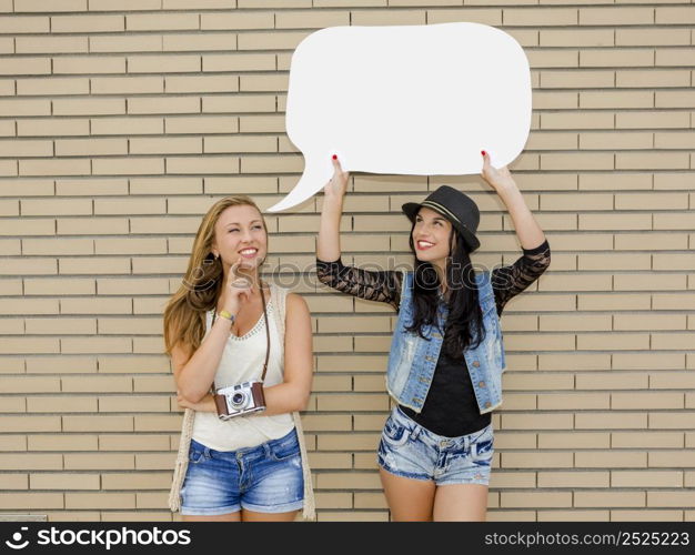 Two beautiful and young girlfriends holding a thought balloon, in front of a brick wall