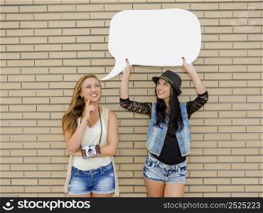 Two beautiful and young girlfriends holding a thought balloon, in front of a brick wall