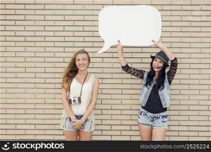 Two beautiful and young girlfriends holding a thought balloon, in front of a brick wall