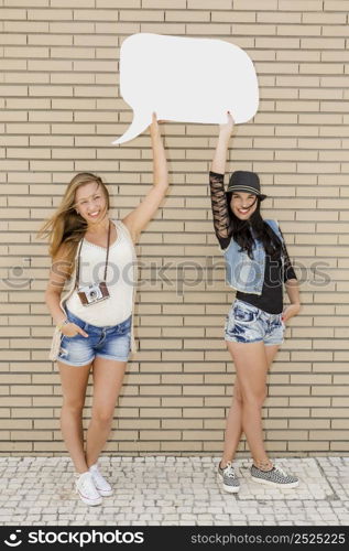 Two beautiful and young girlfriends holding a thought balloon, in front of a brick wall